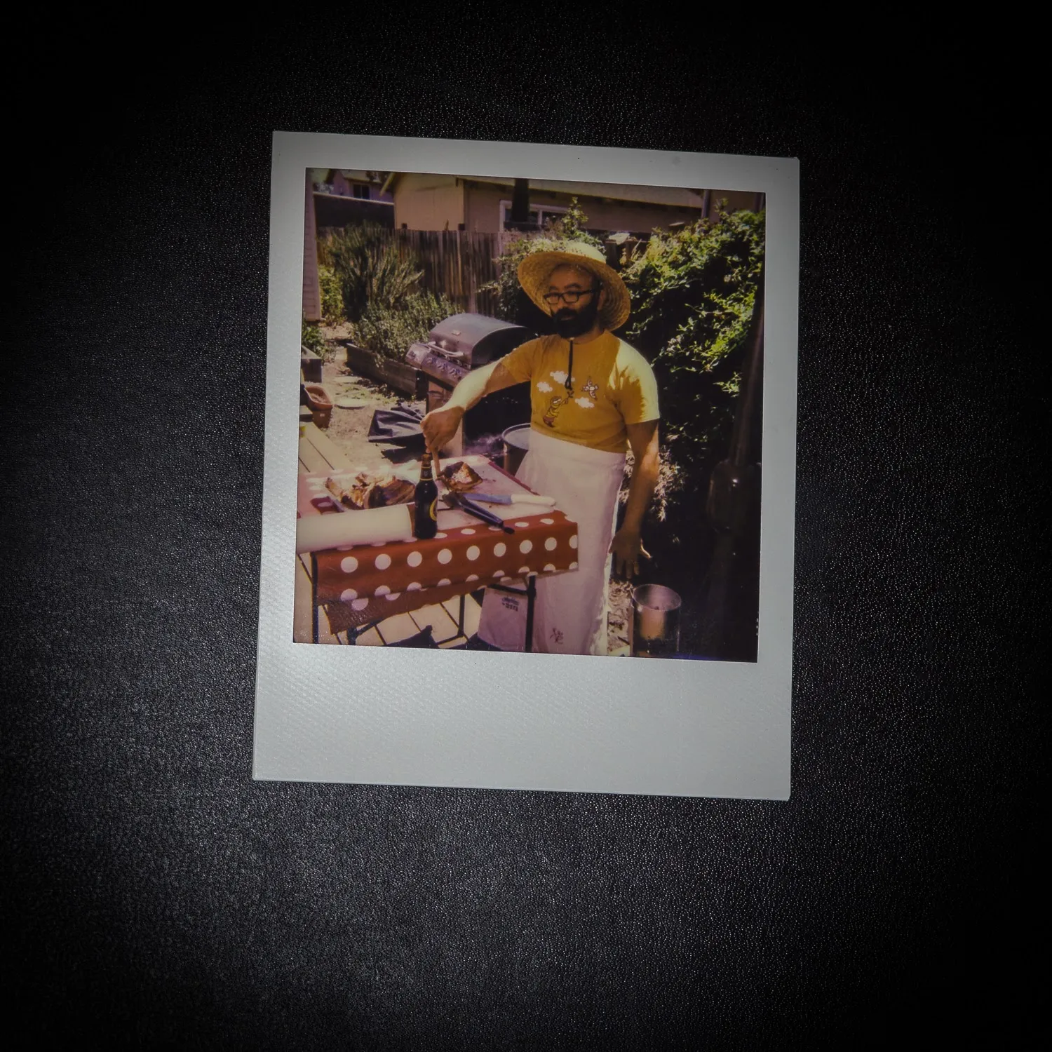 A color print, photographed on black, of a man in a straw hat and apron at a barbecue