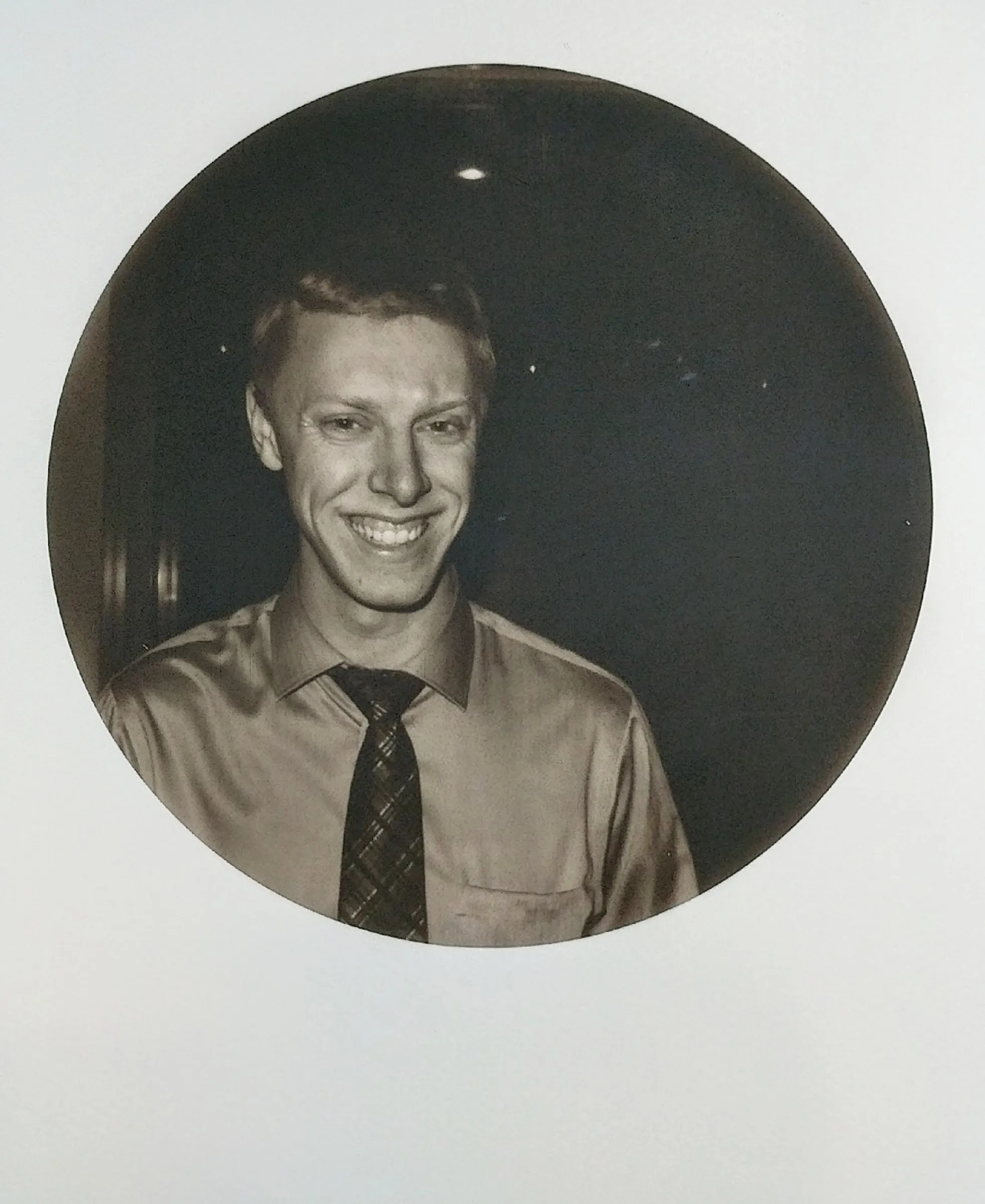 A sepia portrait in a round frame of a young man in a shirt and tie, grinning